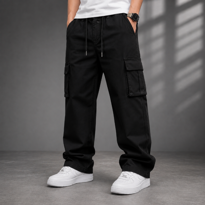 A person wearing black cargo pants with side flap pockets, a white t-shirt, and white sneakers, standing against a gray studio wall with window shadow patterns.