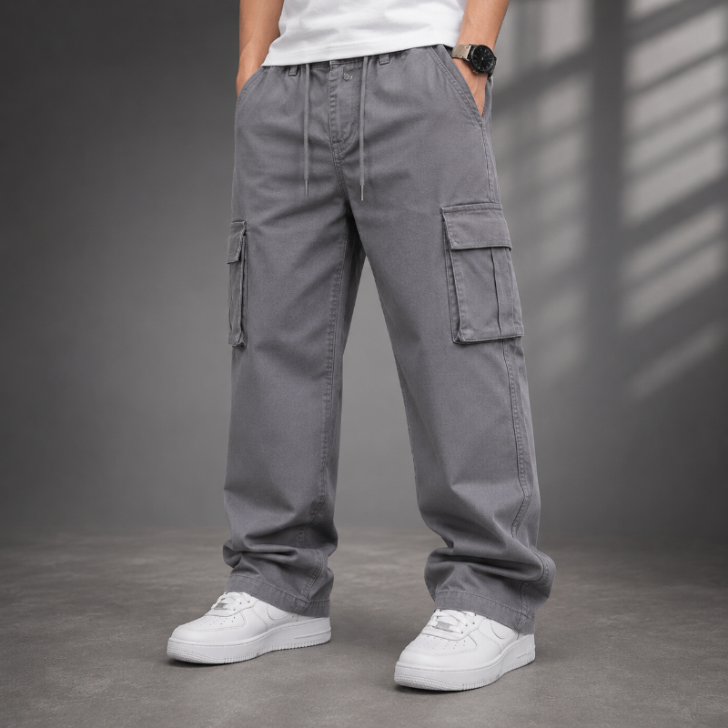 A person wearing loose-fit gray cargo pants with side flap pockets, paired with a white t-shirt and white sneakers, standing against a dark gray studio background with soft window shadow patterns.
