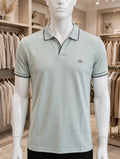 A mannequin torso wearing a light sage green polo shirt with black piping trim on the collar, sleeve cuffs, and a small embroidered logo on the left chest, set inside a clothing store.