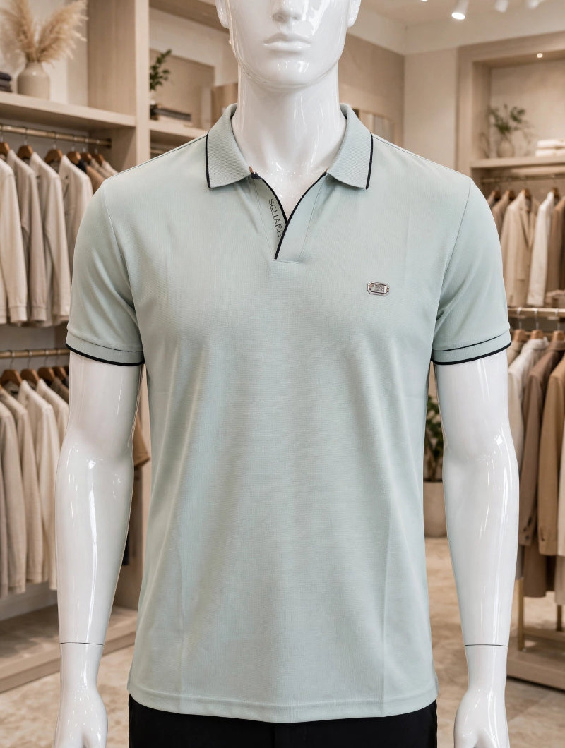 A mannequin torso wearing a light sage green polo shirt with black piping trim on the collar, sleeve cuffs, and a small embroidered logo on the left chest, set inside a clothing store.