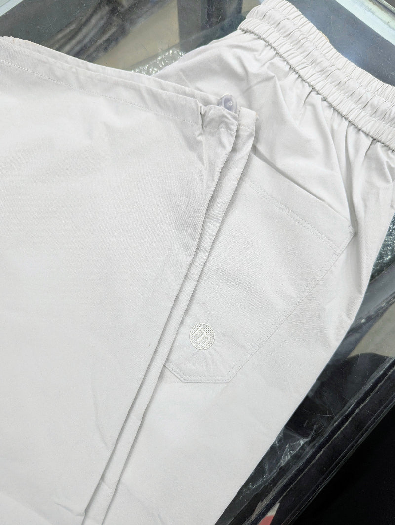 Close-up of a pair of light beige or off-white Baggy Trouser with an elastic waistband and a small embroidered logo on the pocket, laid flat on a dark reflective surface.